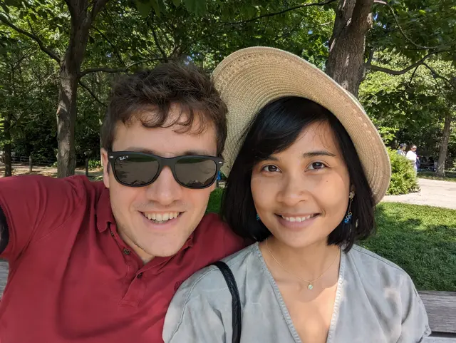 Chris and Emily in a wooded park. Chris has sunglasses on, Emily is wearing a hat.