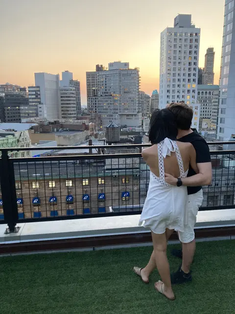 Emily and Chris look off of a rooftop at the sunset. Chris’s hand is on Emily’s back.